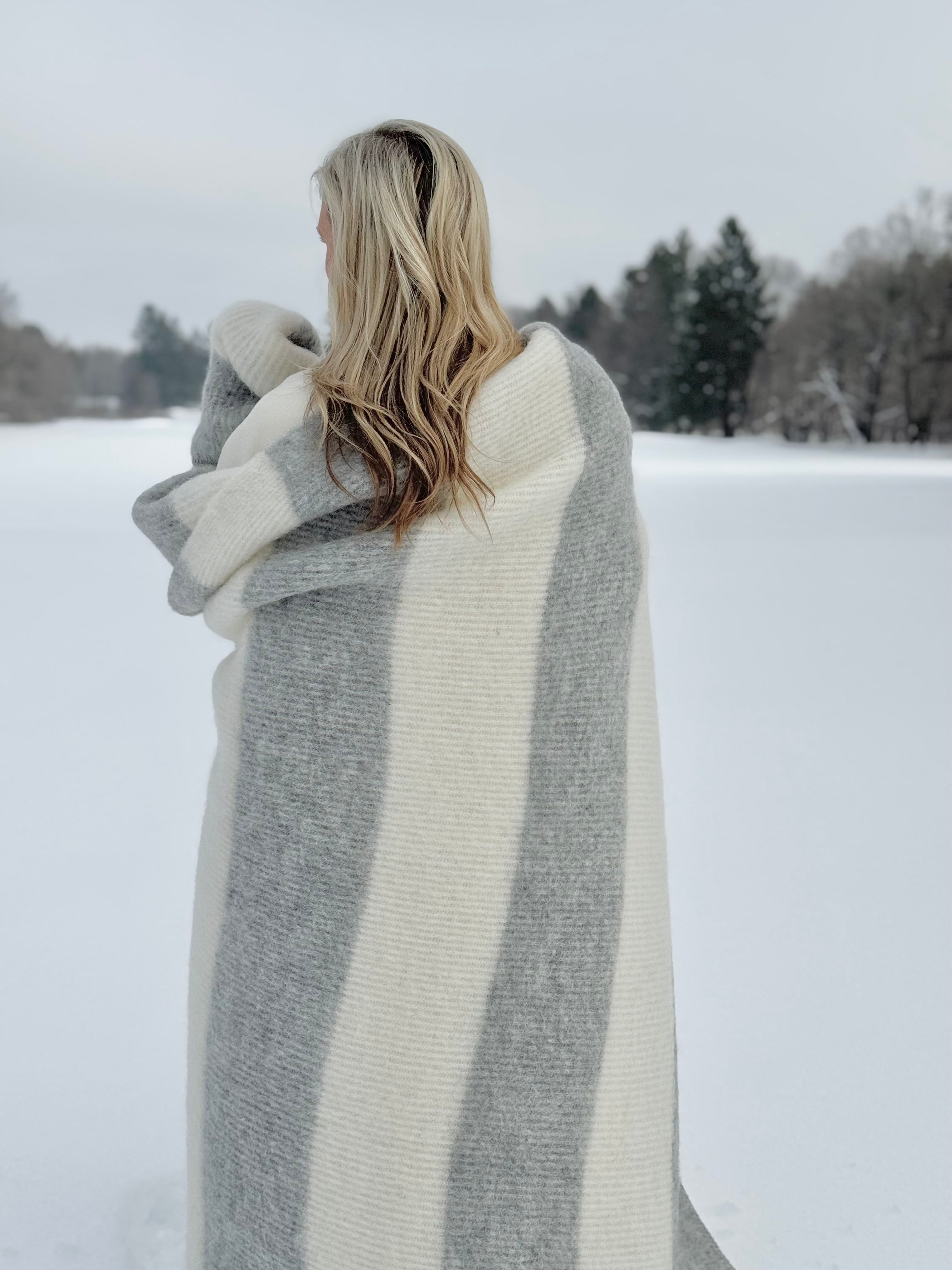 Person wrapped in a large gray and white blanket standing in a snowy landscape.